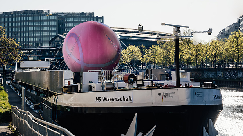 Das Boot "MS Wissenschaft" liegt im Hafen. Auf dem Boot befindet sich eine große pinkfarbene Kugel. Im Hintergrund eine Stadtsilhouette.